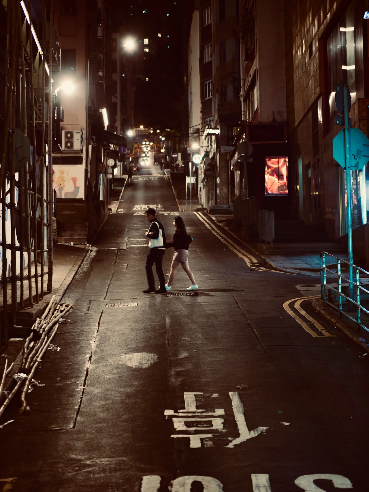 Two people crossing a narrow street under city lights at night.
