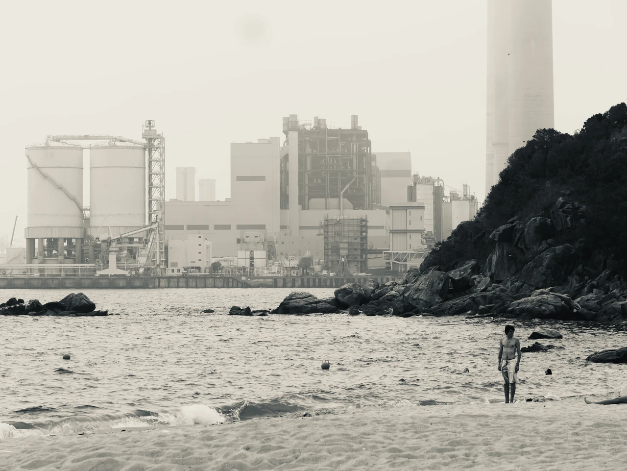A person standing on a beach in front of an industrial waterfront.